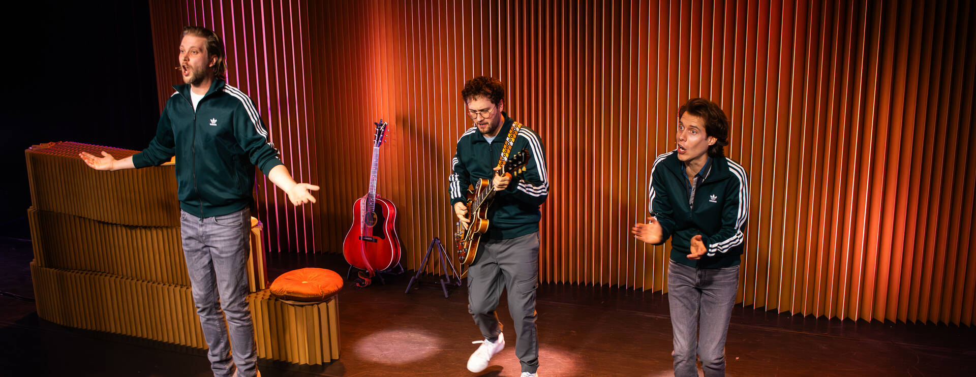 Drie performers in sportoutfits staan op een podium, interactief met het publiek en een gitaar op de achtergrond.
