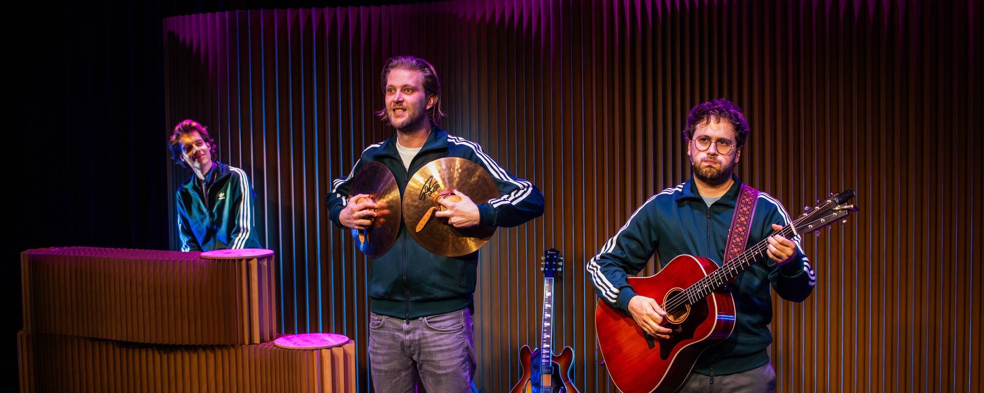 Drie muzikanten op een podium; een speelt keyboard, terwijl de anderen een gitaar en percussie bespelen.