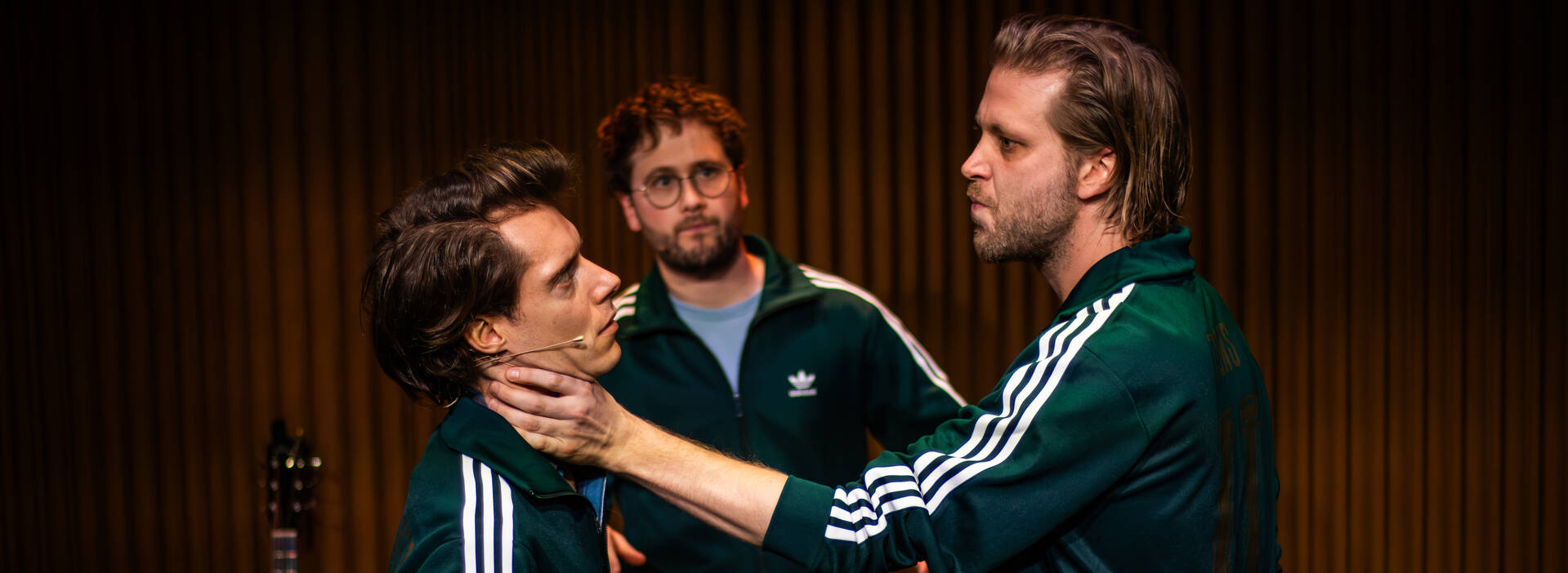 Drie acteurs in sportoutfits, met één acteur die de ander vasthoudt bij de keel in een intense scène.