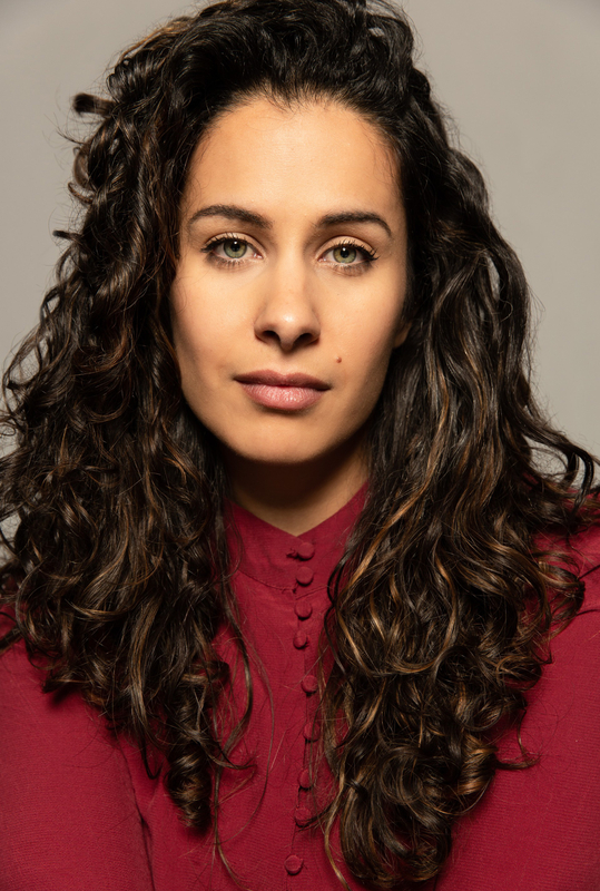 Portret van een vrouw met krullend bruin haar, gekleed in een rode blouse, recht in de camera kijkend.