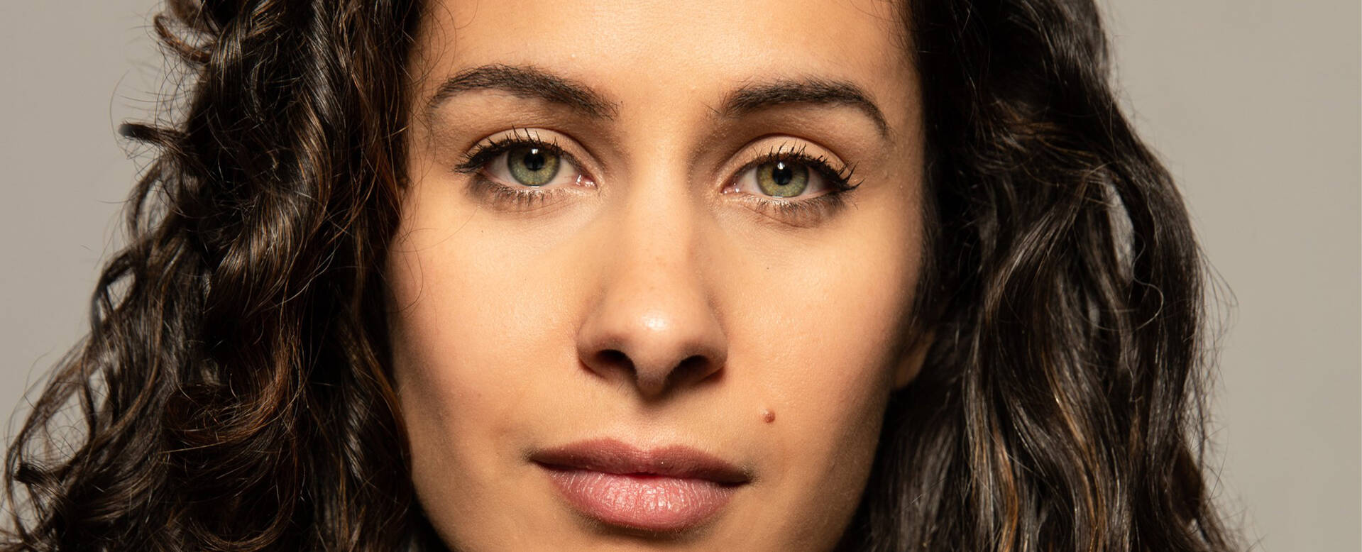 Portret van een vrouw met krullend bruin haar en een neutrale gezichtsuitdrukking op een effen achtergrond.