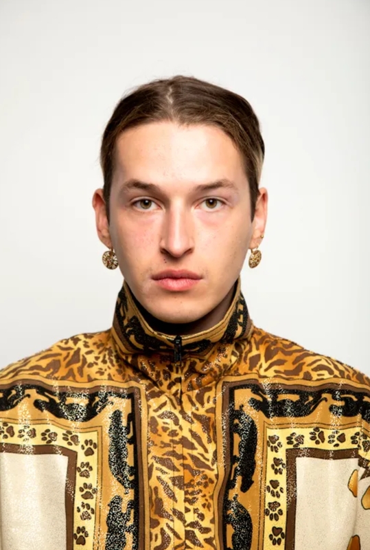 Portret van een jonge man met een opvallende jas met dierenprint en grote oorbellen, tegen een neutrale achtergrond.