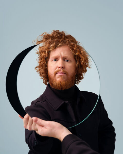 Man met krullend haar en baard houdt een halfronde spiegel voor zich in een lichtblauwe achtergrond.