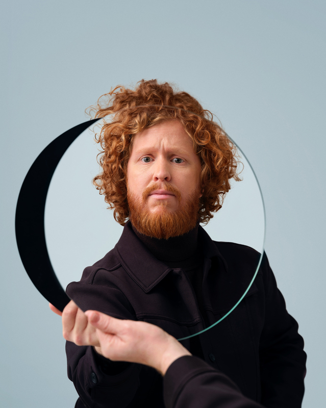 Man met krullend haar en baard houdt een halfronde spiegel voor zich in een lichtblauwe achtergrond.