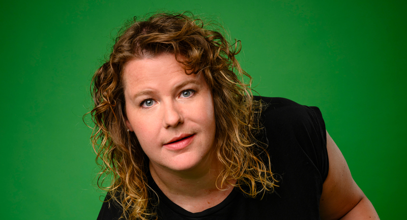 Portret van een vrouw met krullend haar, die met een neutrale gezichtsuitdrukking naar de camera kijkt tegen een groene achtergrond.