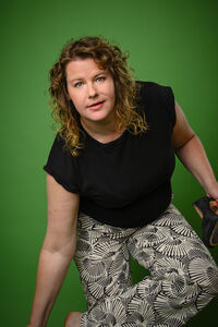 Vrouw met krullend haar in zwarte shirt en broek met motief, zittend op een knielende positie tegen een groene achtergrond.