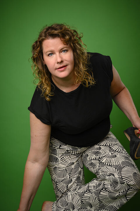 Vrouw met krullend haar in zwarte shirt en broek met motief, zittend op een knielende positie tegen een groene achtergrond.