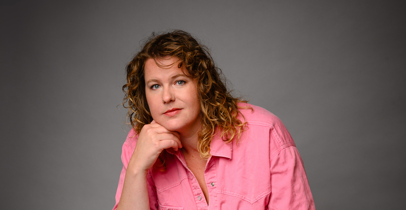 Vrouw met krullend haar in een roze blouse, leunend op haar hand tegen een neutrale achtergrond.