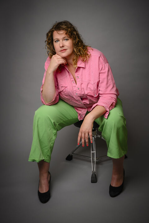 Vrouw met krullend haar in roze blouse en groene broek, zittend op een stoel met een neutrale achtergrond.