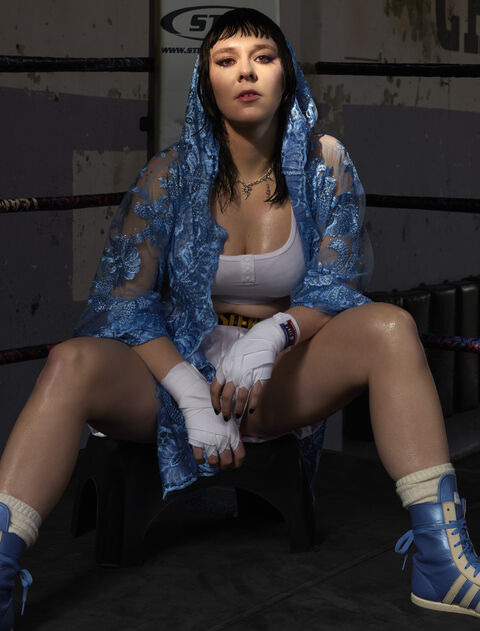 Vrouw in boksring in een blauwe outfit, zittend met ingepakte handen en een zelfverzekerde houding.