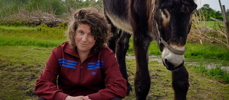Een vrouw zit op de grond naast een zwart paard in een groene, open omgeving.