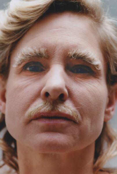 Portret van een persoon met een neutrale uitdrukking, lichte krullende haren en een blond snor.