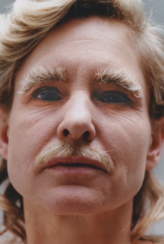 Portret van een persoon met een neutrale uitdrukking, lichte krullende haren en een blond snor.