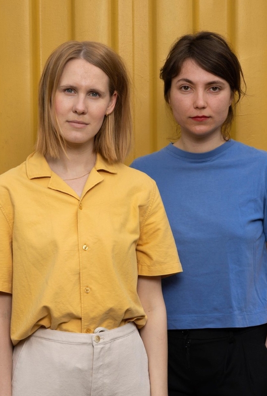 Twee vrouwen staan naast elkaar voor een gele achtergrond, met neutrale gezichten en casual kleding.