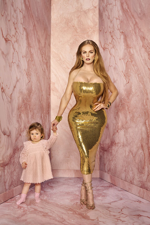 Een vrouw in een gouden, strakke jurk staat samen met een klein meisje in een roze marmeren omgeving.