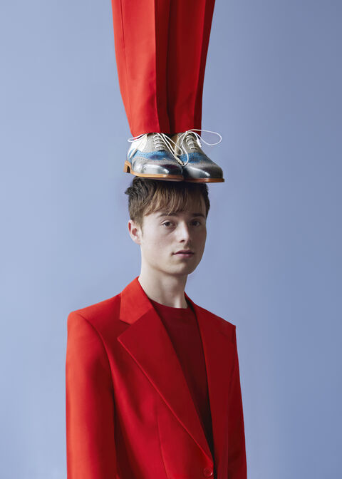 Jongeman in een rood pak met een paar schoenen op zijn hoofd, tegen een blauwe achtergrond.