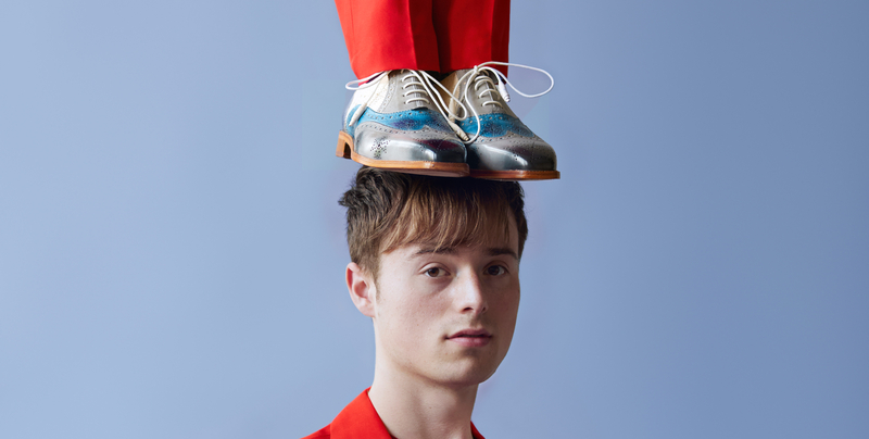 Jongeman met een neutrale uitdrukking, met schoenen op zijn hoofd, gekleed in een felle rode outfit tegen een blauwe achtergrond.