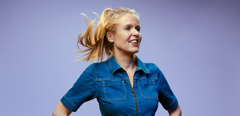 Vrouw met een glimlach en een hoge paardenstaart, gekleed in een blauwe jurk, tegen een paarse achtergrond.