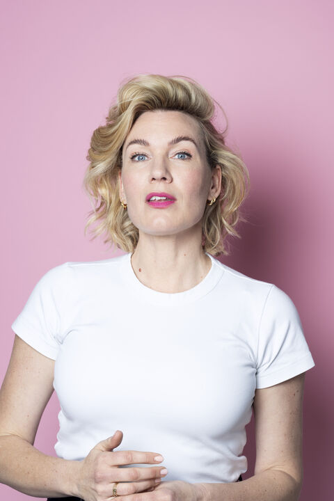 Portret van een vrouw met een lichte huid en blond, krullend haar, staand voor een roze achtergrond. Ze kijkt recht in de camera.