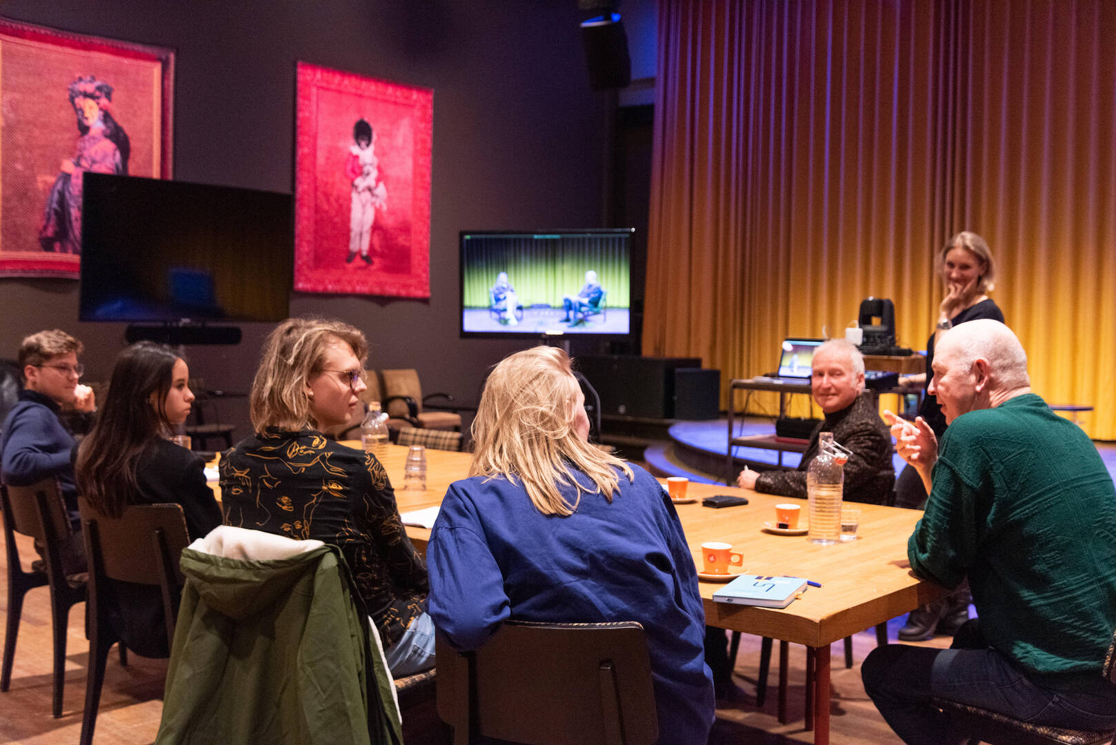 Groep mensen in een zaal, zittend aan een tafel, met kunstwerken en een presentatie op de achtergrond.