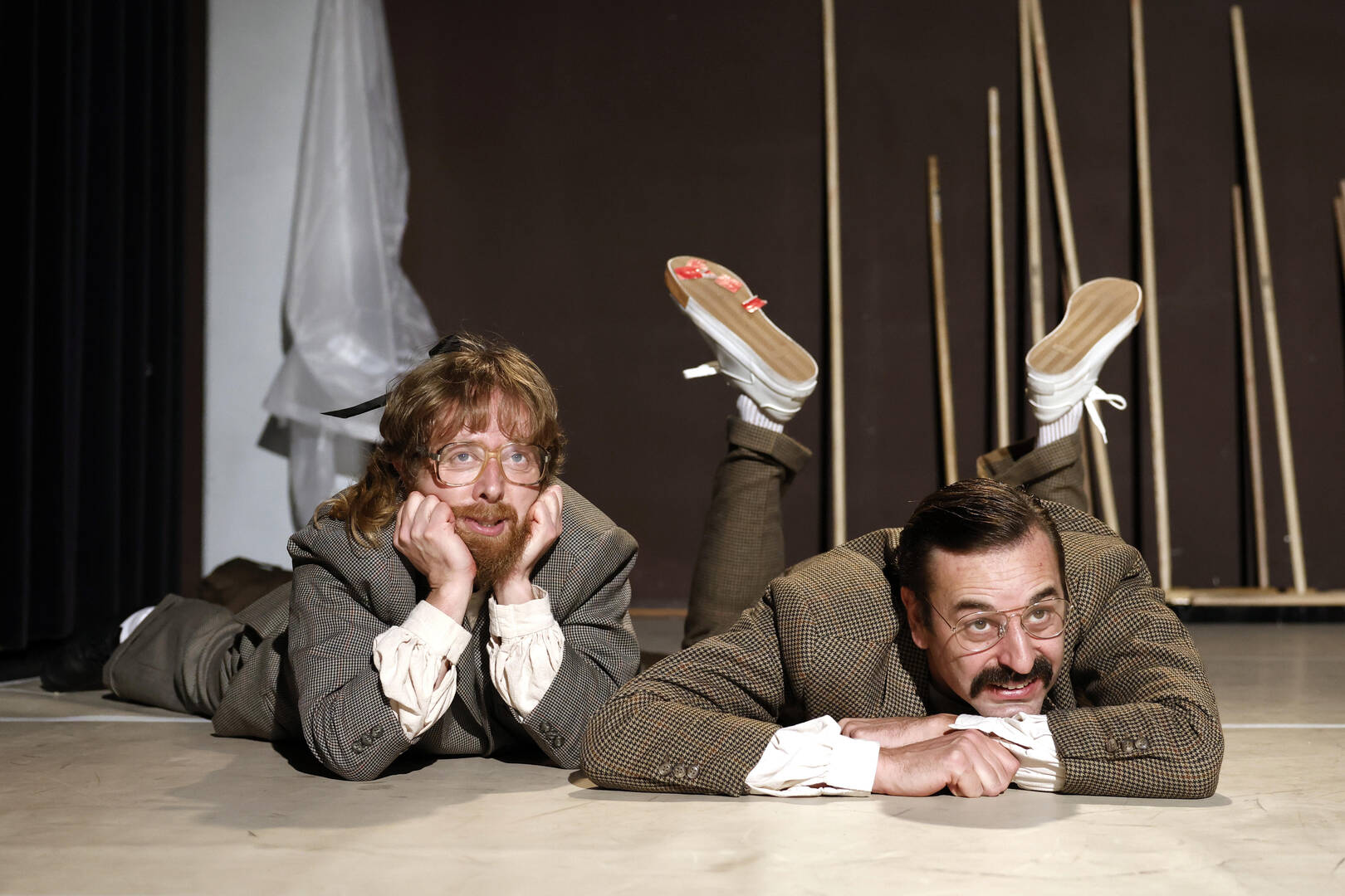 Twee mannen in historische kostuums liggen op de vloer van een toneel met een speelse houding.