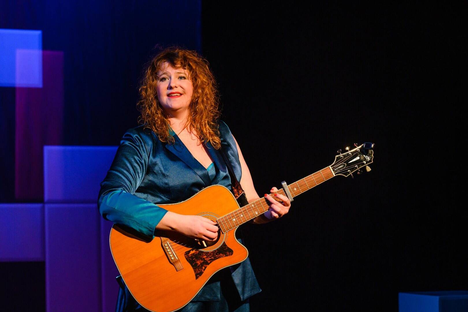 Vrouw met krullend haar in een blauwe outfit speelt gitaar op een podium met kleurrijke achtergrondverlichting.