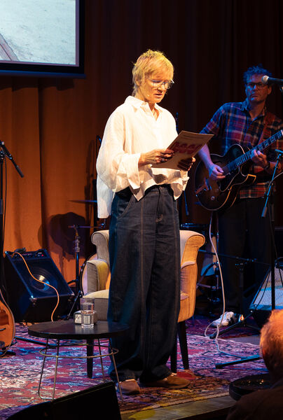 Een vrouw met een boek in de hand staat op een podium met een stoel en muzikanten in de achtergrond.
