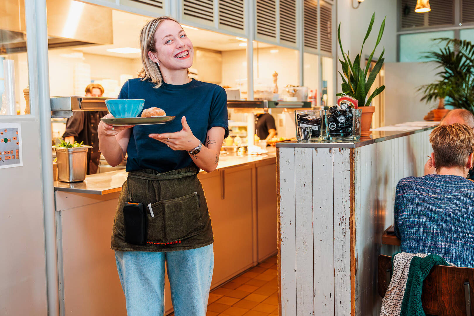 Vrouw in een restaurant serveert een schotel met een blauwe kom, glimlachend naar de camera.