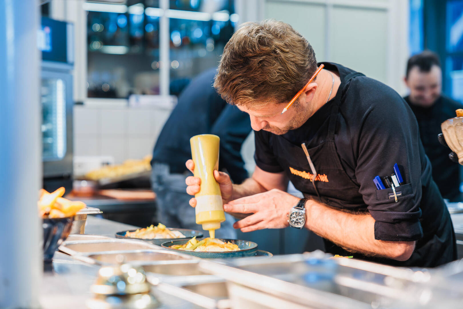 Een kok garneert een gerecht met een gele saus uit een knijpfles in een keuken.