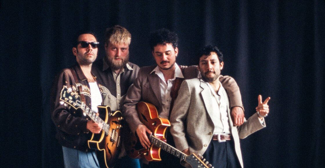 Vier muzikanten poseren samen met gitaren, gekleed in retro-stijl outfits voor een donkere achtergrond.