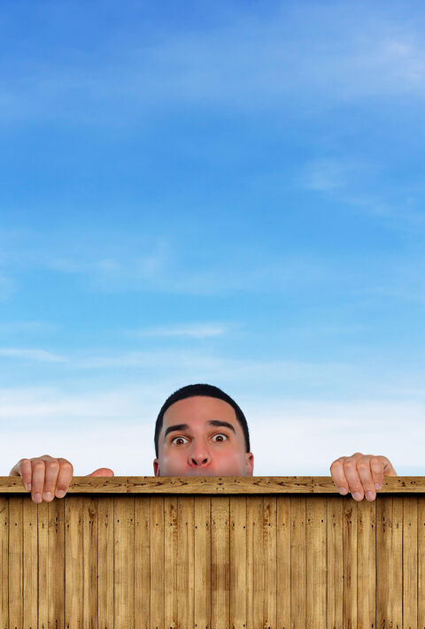 Man met een verbaasde uitdrukking kijkt over een houten schutting onder een blauwe lucht.