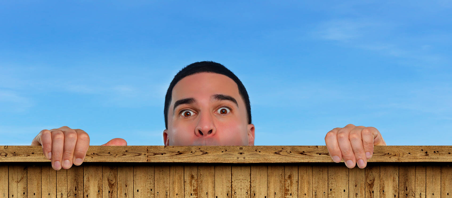 Een man kijkt nieuwsgierig boven een houten hek uit met een blauwe lucht op de achtergrond.
