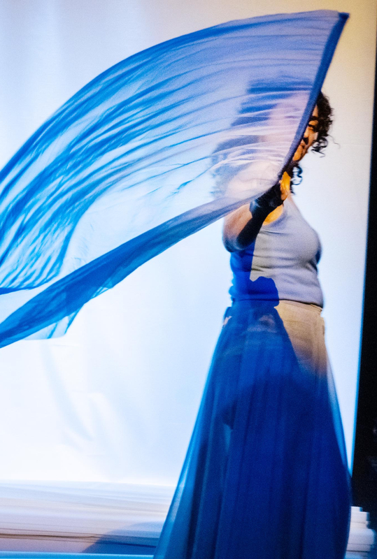 Vrouw in een blauw top en rok, die een doorschijnende blauwe sjaal of doek laat wapperen in een dansachtige pose.