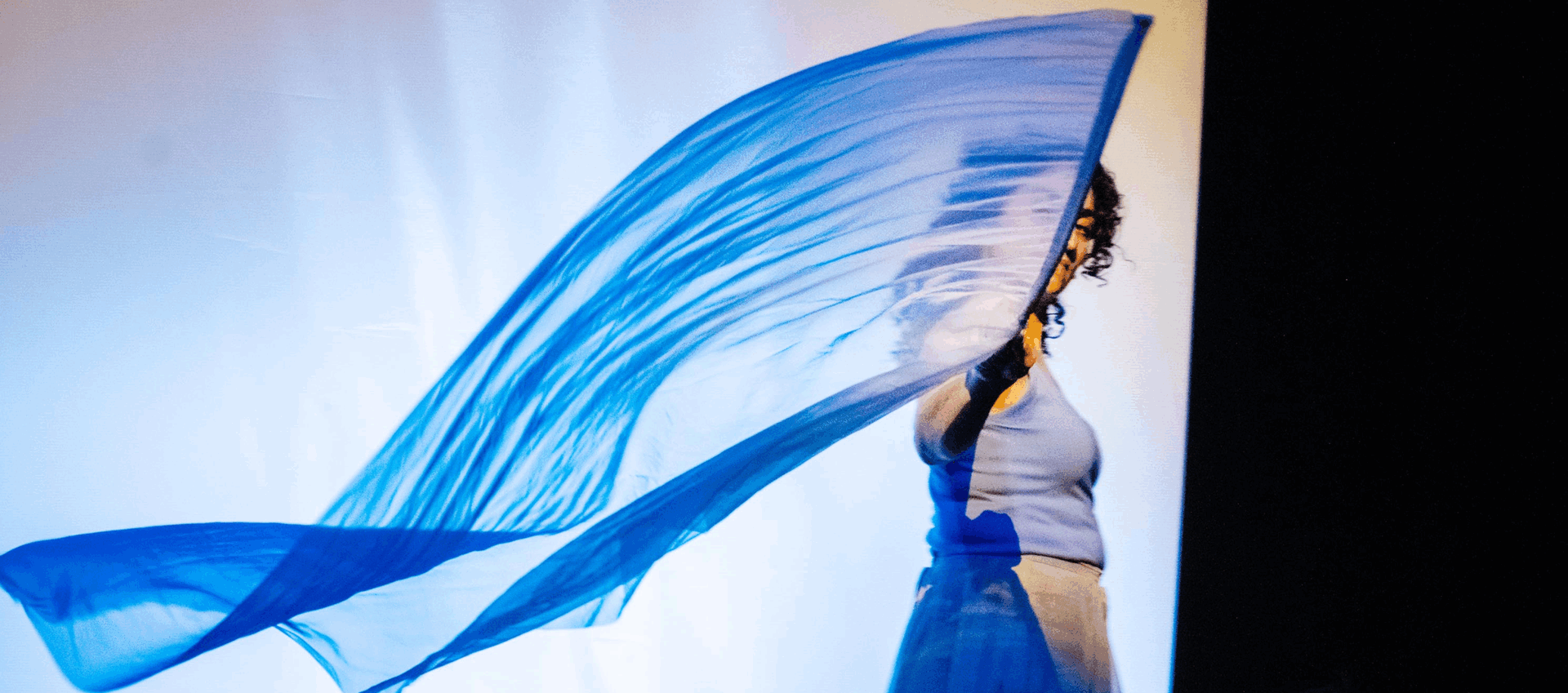 Vrouw in blauw kostuum die een grote blauwe doek omhoog houdt, tegen een lichte achtergrond.