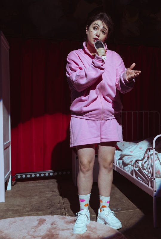 Een vrouw in een roze outfit zingt in een uitdagende setting met een bed op de achtergrond.