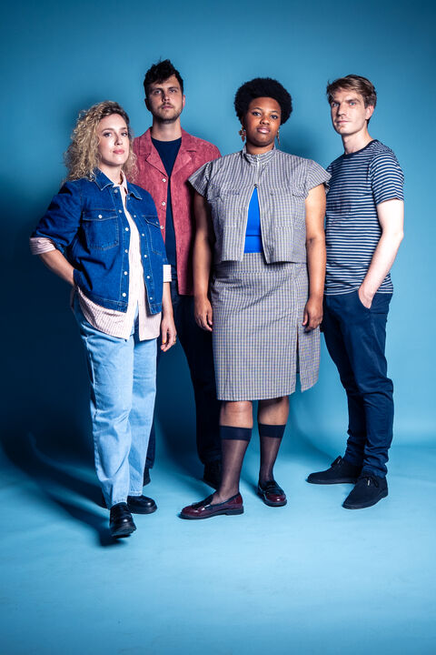 Groep van vier mensen staande tegen een blauwe achtergrond, gekleed in casual en moderne kleding.