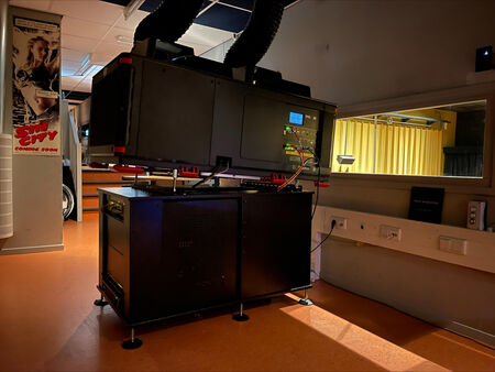 Zwart apparaat op een bureau in een ruimte met een venster en apparatuur tegen de muur. Achtergrond met gele gordijnen.