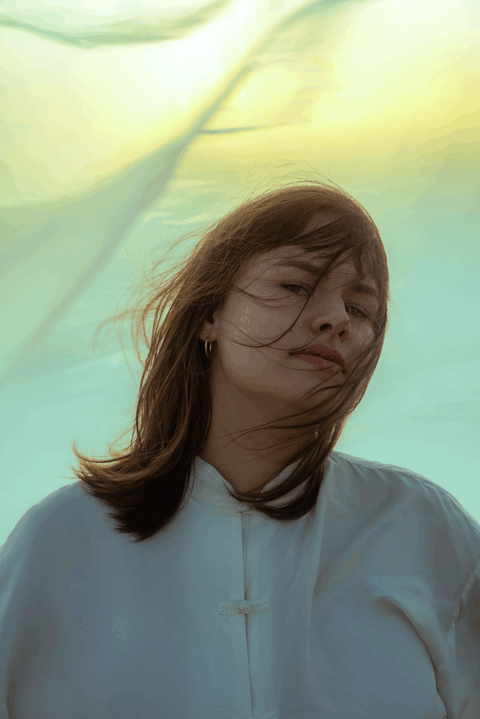 Portret van een vrouw met winderig haar, gekleed in een witte blouse, met een kleurrijke achtergrond van bewegende stof.