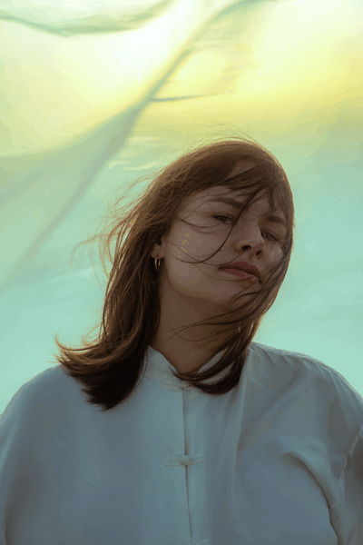 Portret van een vrouw met winderig haar, gekleed in een witte blouse, met een kleurrijke achtergrond van bewegende stof.