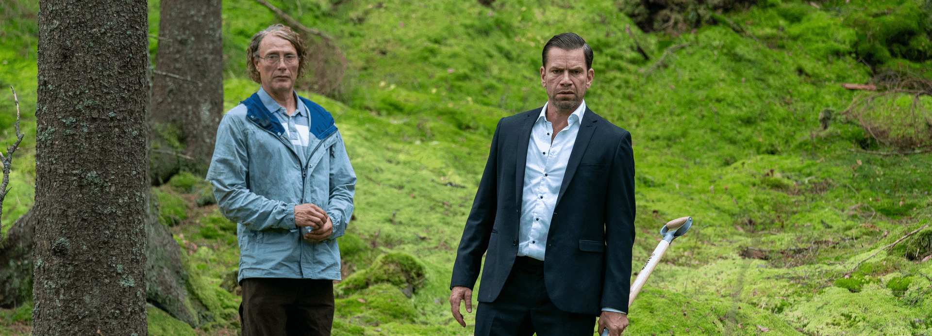 Twee mannen staan in een bosrijke omgeving, omringd door groene begroeiing. Eén in casual kleding, de ander in een pak.
