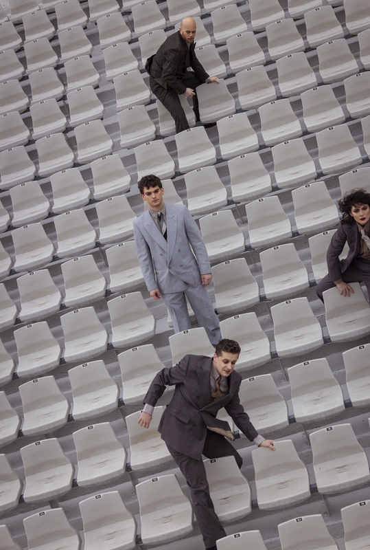 Groep mannen in maatpakken die op een trap van lege stoelen staan, met één man die achteraan loopt.