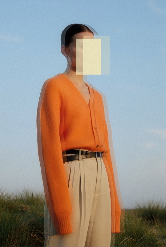 Vrouw in een oranje trui en beige broek, met een onduidelijke gezichtsweergave, staand tegen een blauwe lucht.