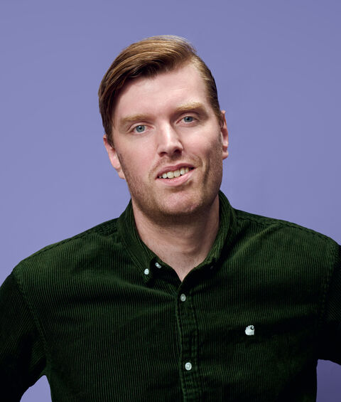 Portret van een man met kort, bruin haar in een groene blouse, staand tegen een paarse achtergrond.