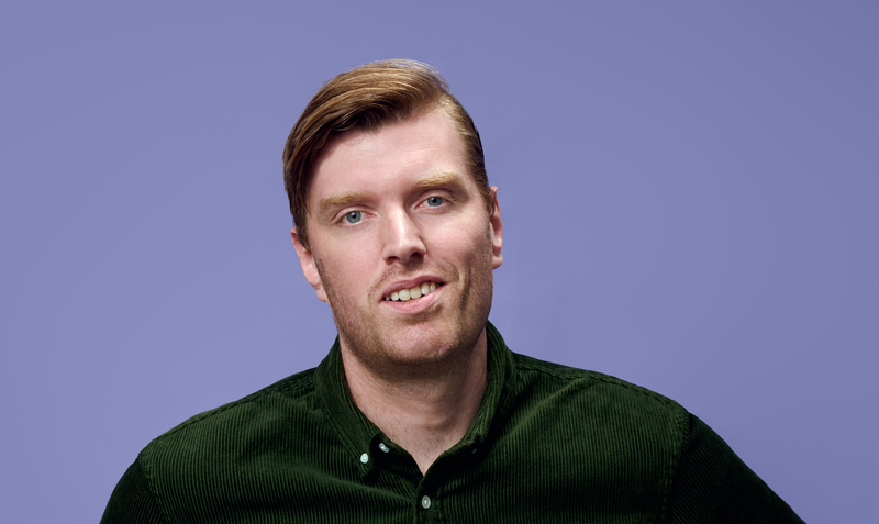 Man met een lichte glimlach en bruin haar, staande voor een paarse achtergrond. Draagt een groene shirt.