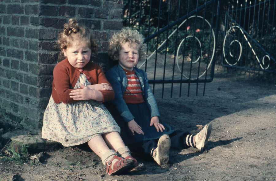 Twee kinderen met krullend haar zitten op de grond bij een muurtje, omgeven door een hek en groene omgeving.