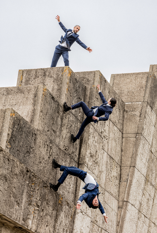 Drie personen in pak poseren op een architectonische betonstructuur, met een dynamische houding van vallen en stijgen.