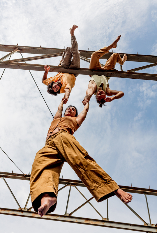 Drie acrobaten hangend aan een metalen constructie, één ondersteund door de anderen, met een heldere lucht op de achtergrond.
