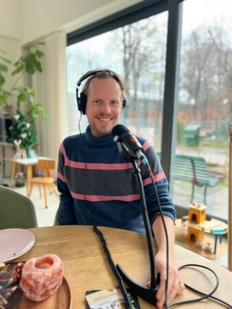 Mannelijke podcaster met koptelefoon zit aan tafel met microfoon, omringd door een lichte en moderne inrichting.