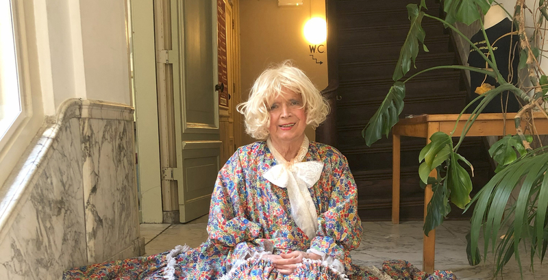 Vrouw met een blonde pruik in een kleurrijk kledingstuk, zittend in een lichte, moderne ruimte.