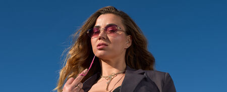 Vrouw met lange, golvende haren in een zonnige omgeving, draagt een donkergrijze blazer en roze zonnebril.